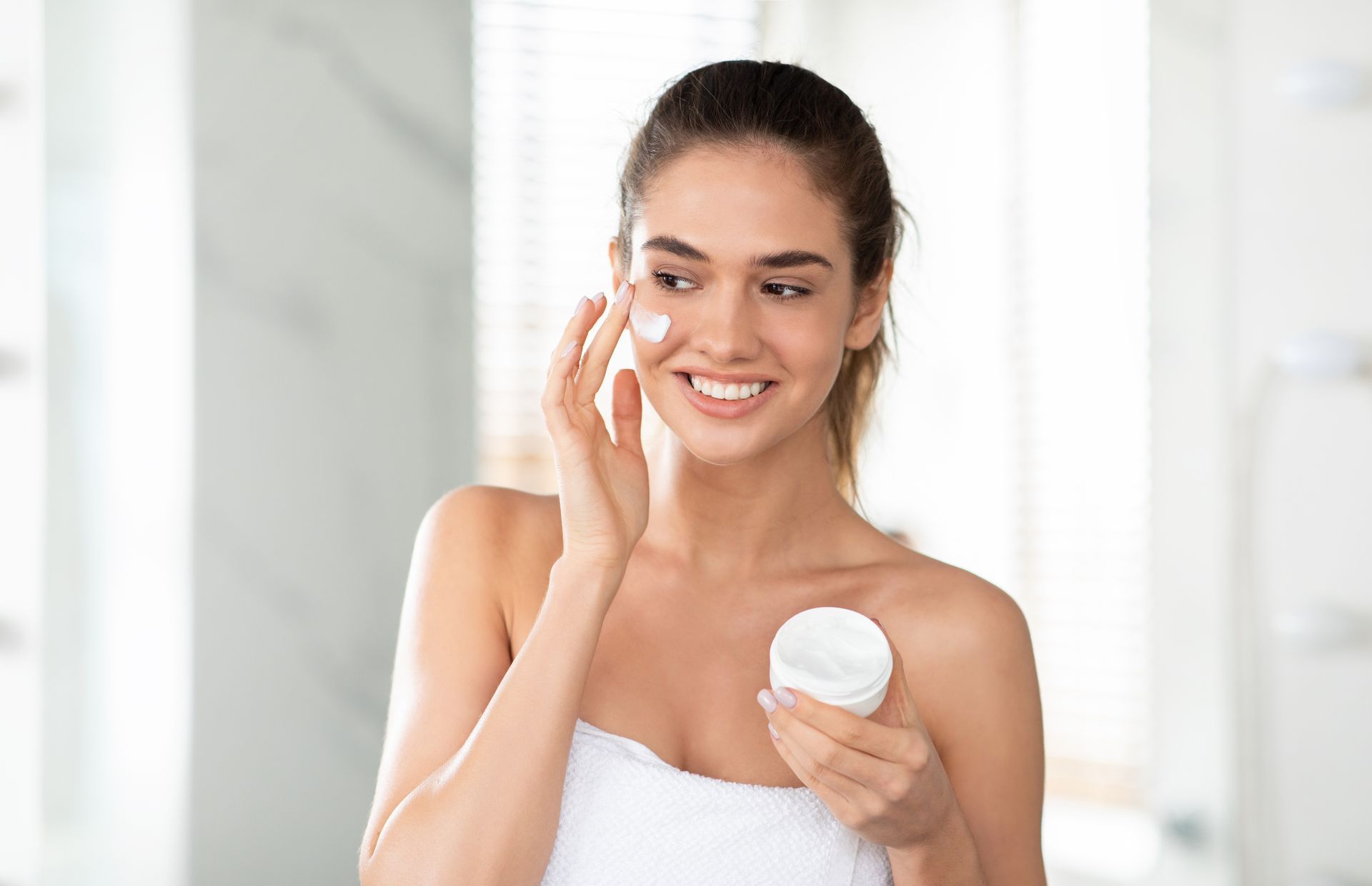 A woman wrapped in a towel is applying cream to her face.