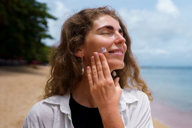 A woman is applying sunscreen to her face on the beach.