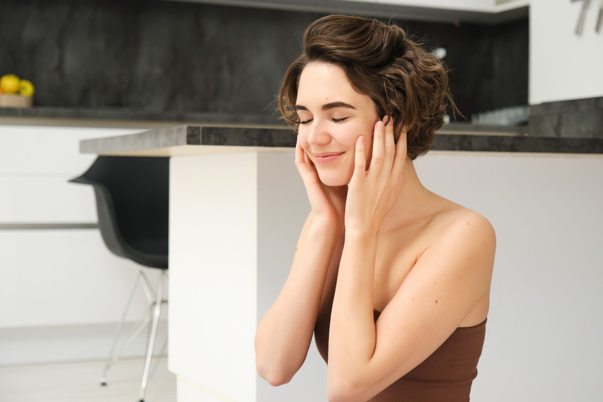 A woman is sitting in a kitchen with her eyes closed and her hands on her face.