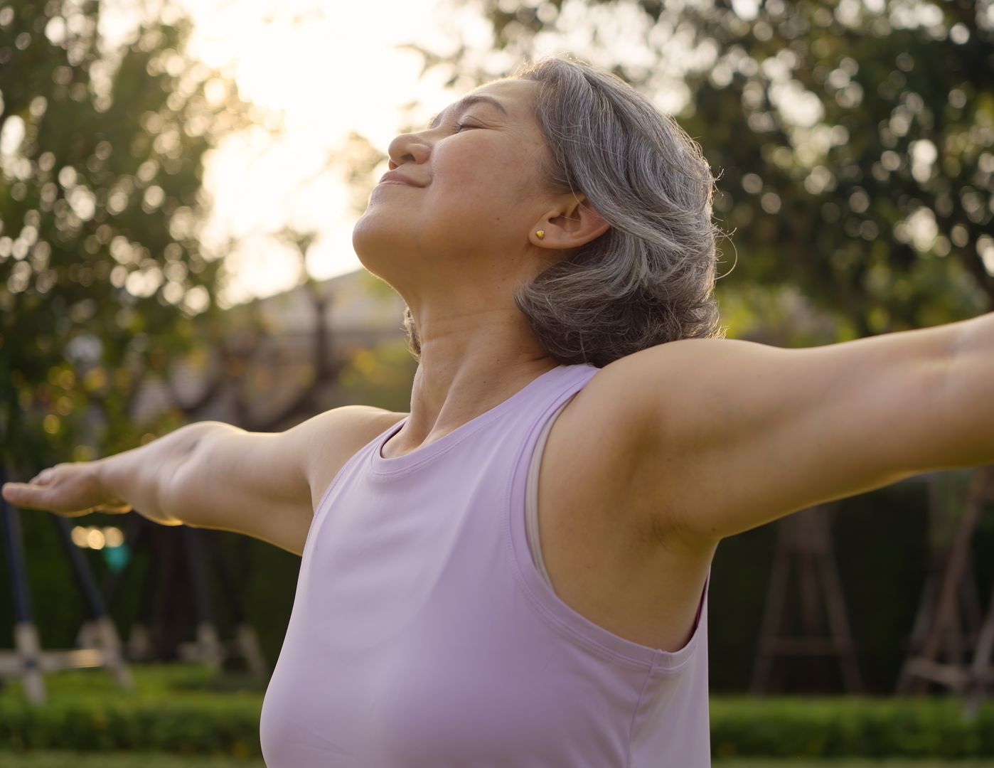 Woman in a lavender top with arms outstretched, smiling in a sunlit park.