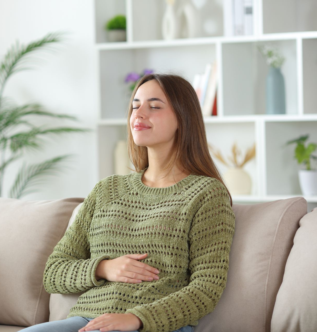 Woman relaxing on a couch with eyes closed, wearing a green sweater in a bright living room