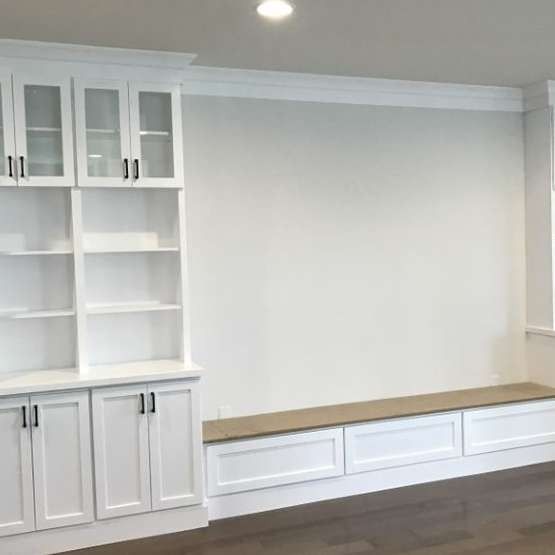 White built-in shelving unit with glass-front cabinets and a window seat in a room with light-colored walls.