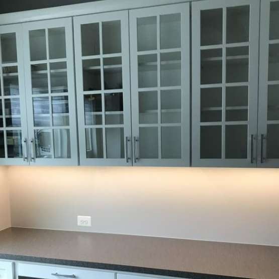 Light gray kitchen cabinets with glass pane doors over a dark countertop with under-cabinet lighting.