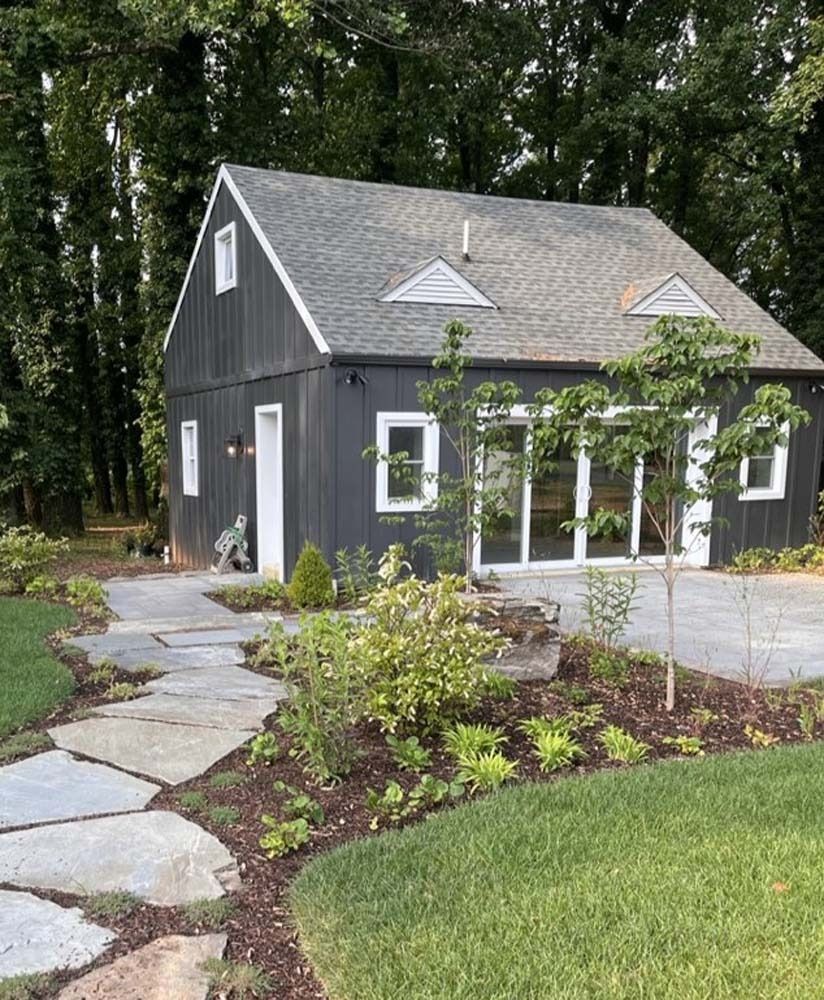 Charcoal-colored barn-style building with stone path and landscaping; trees in the background.