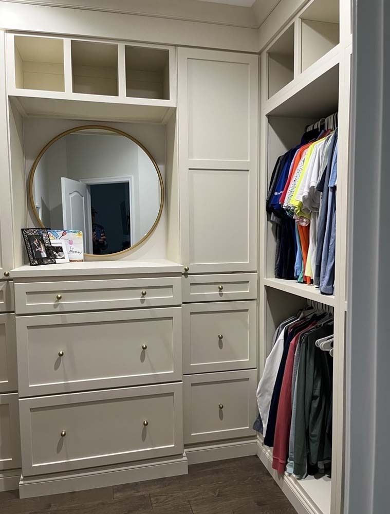 Beige custom closet with drawers, shelves, hanging rods, and a round mirror.
