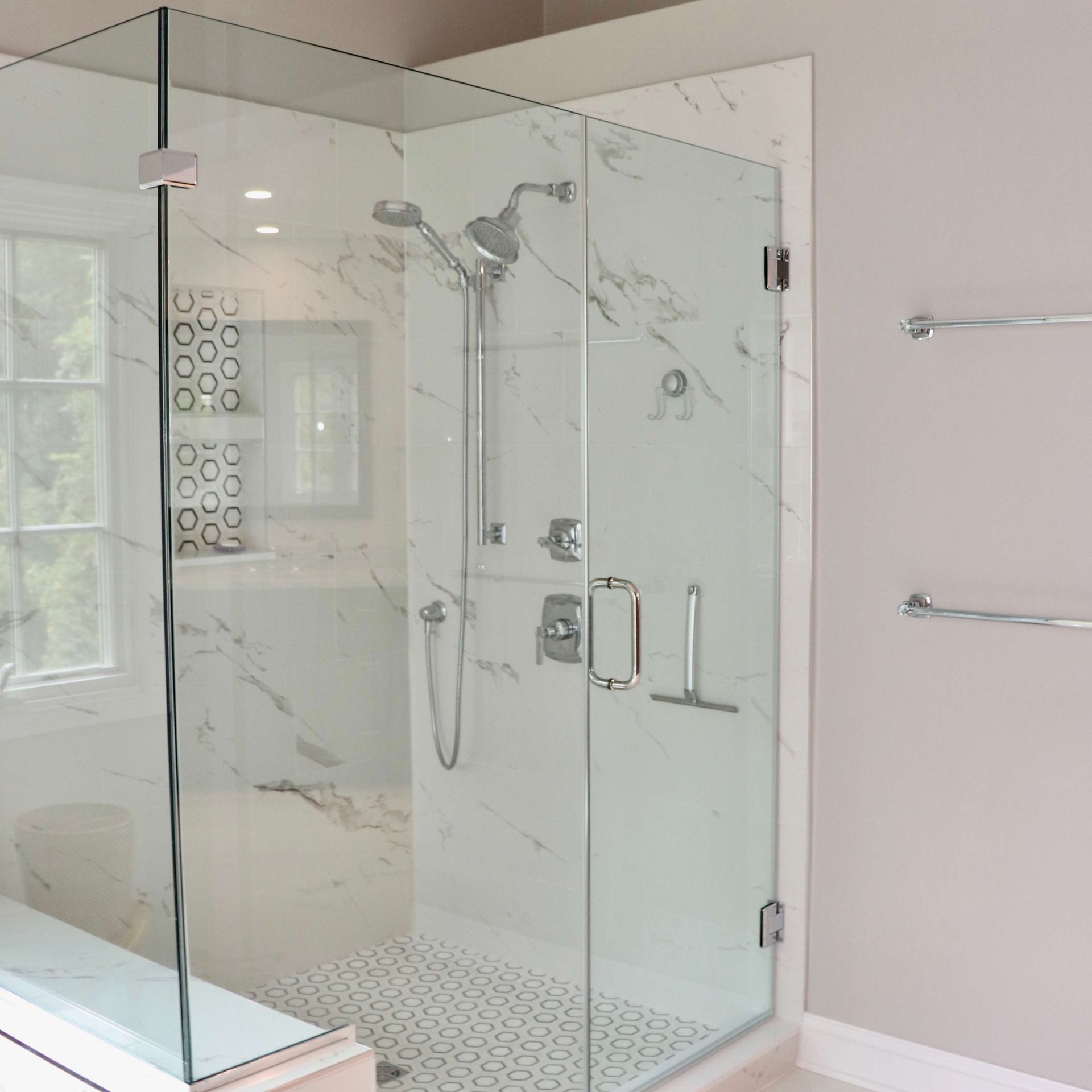 Glass-enclosed shower in a bathroom with marble walls, chrome fixtures, and a hexagonal tiled floor.