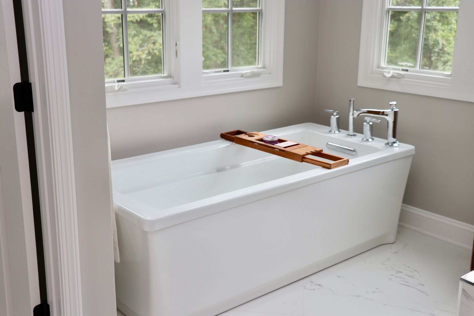 White bathtub in a bathroom with windows; wooden bath tray, chrome faucet.