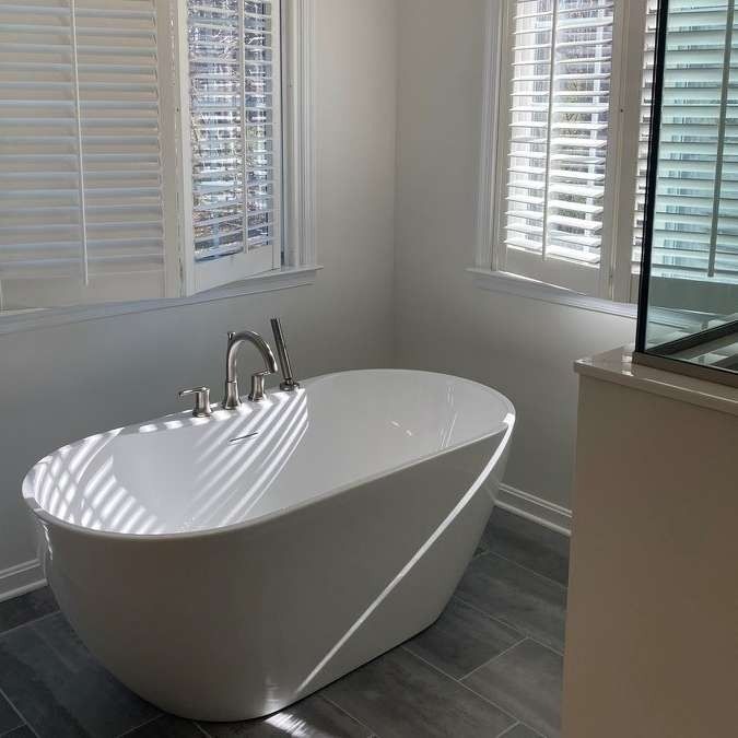 White oval bathtub in a bright bathroom, next to windows with white shutters.