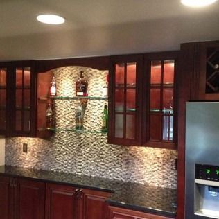 A dark wood home bar with a granite countertop, glass shelves, and lit cabinets.