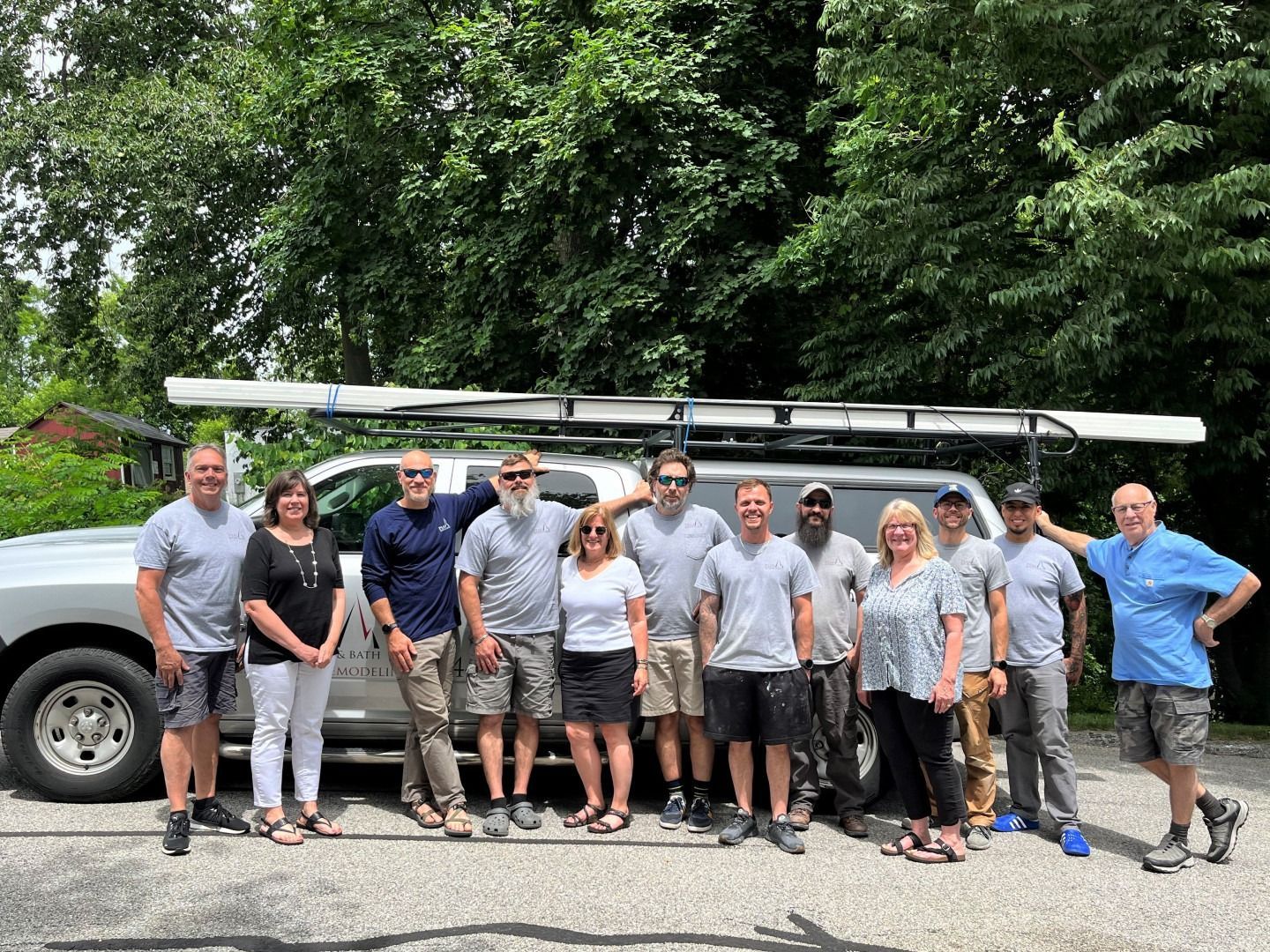 Group of people standing in front of a truck with oars on top, outdoors, in front of trees and a building.