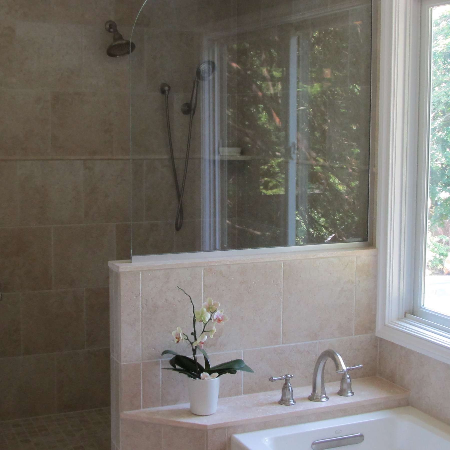 Bathroom with beige tiled shower, bathtub, and window with outdoor view. White flower in a pot on the ledge.