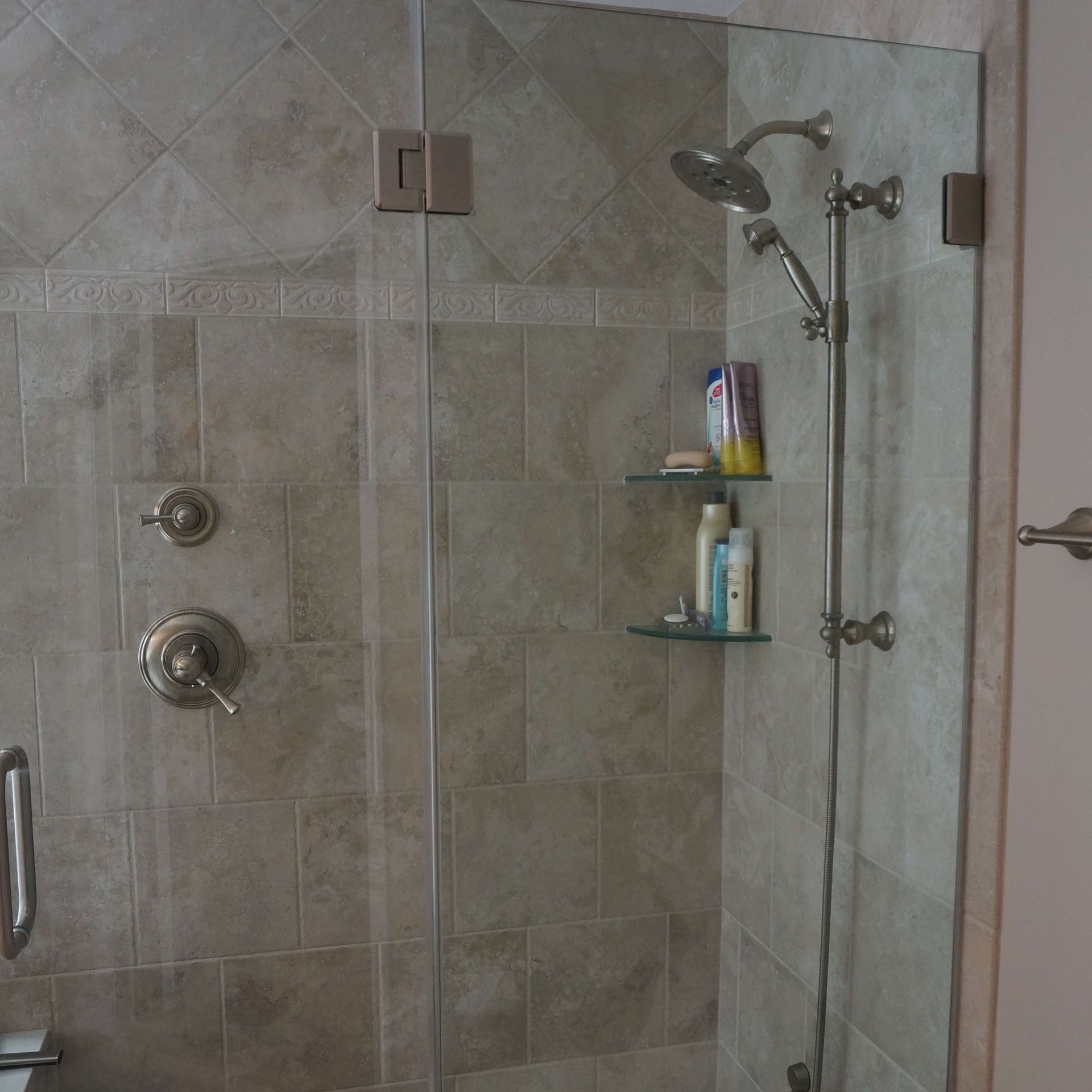 Glass-enclosed shower with beige tile walls. Showerhead, shelves with products, and fixtures.