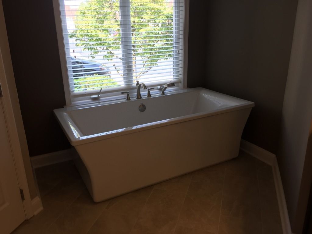Freestanding white bathtub under a window with blinds in a bathroom.