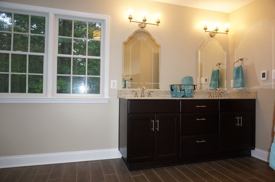Bathroom with dark brown vanity, two arched mirrors, and a window. Beige walls, hardwood floor, and teal accents.