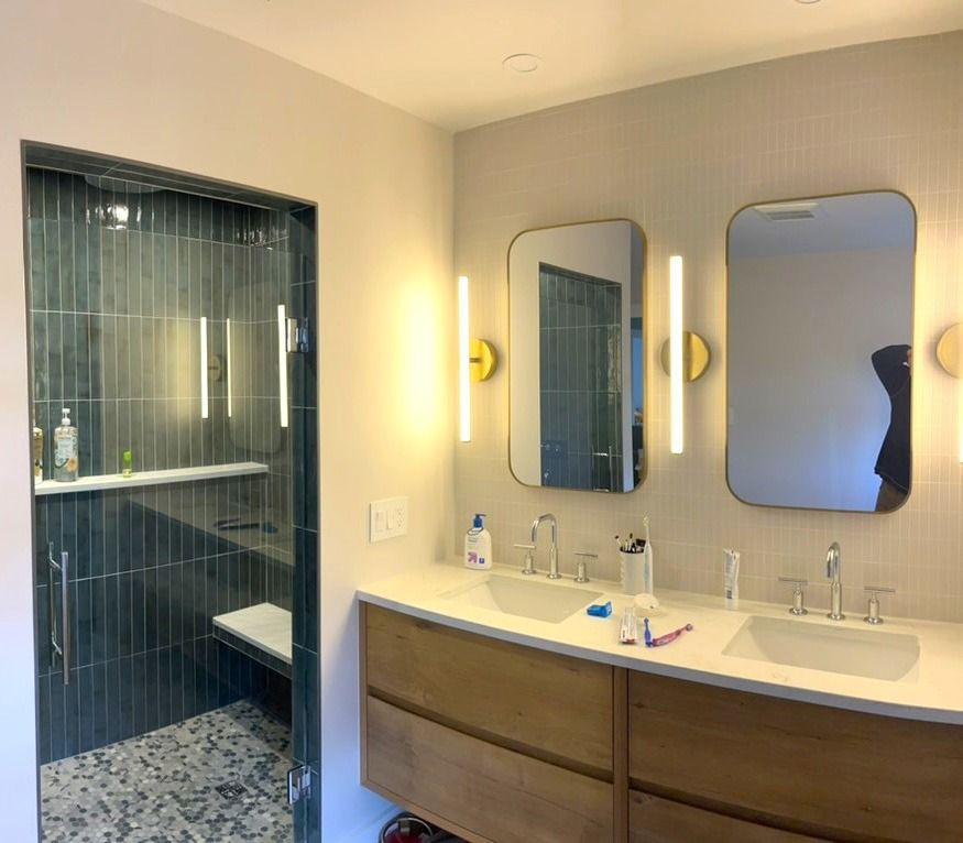 Bathroom with double sinks, mirrors, shower with dark tile, and light wood vanity.