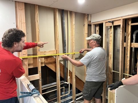 Two men measure a wall during construction; one points, other holds tape. Exposed framing, pipes visible.