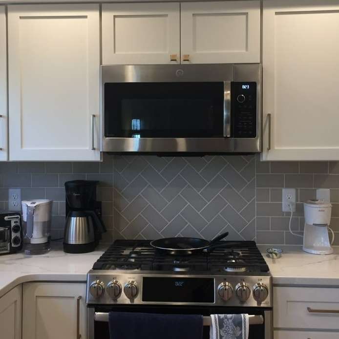 A kitchen with white cabinets, a gray herringbone backsplash, stainless steel microwave, and a stove with a pan.