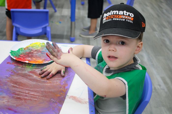 A young boy wearing a holmatro hat is sitting at a table with his hands covered in paint.