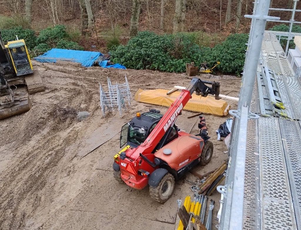 Rød teleskoplæsser på mudret jord nær stilladser og træer.