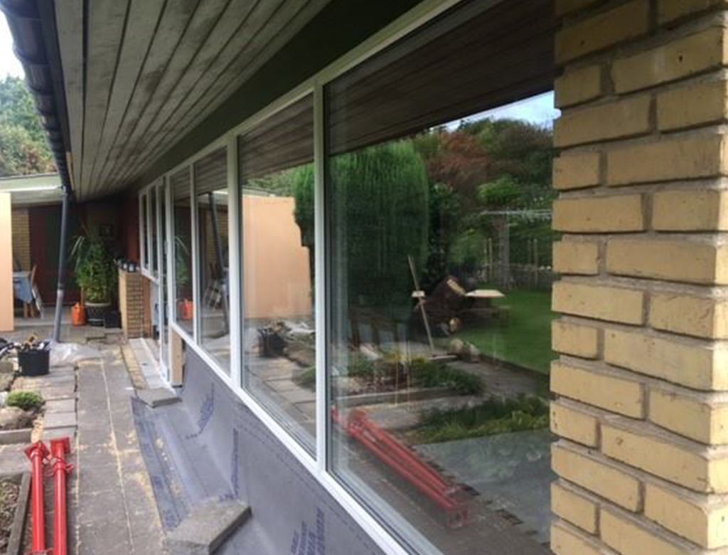 Udvendig udsigt af huset med store vinduer, der afspejler en have. Murstensvæg til højre, træbeklædning over vinduerne.