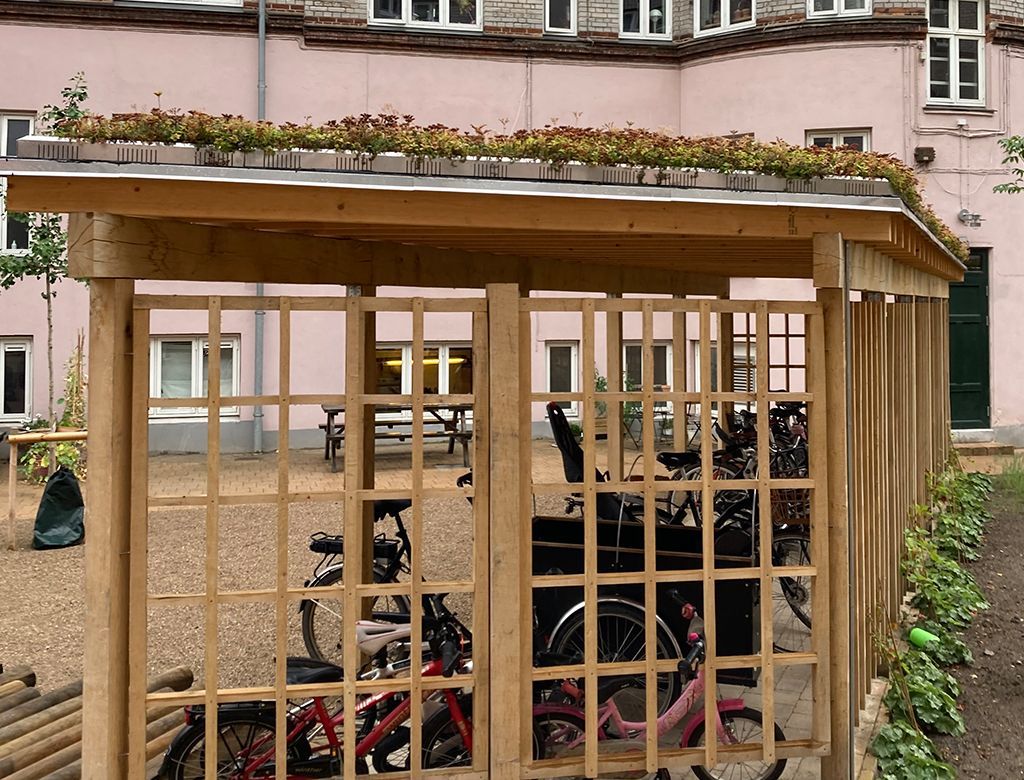 Cykelskur i træ med gitterpaneler på siderne og grønt tag, cykler indenfor, foran en bygning.