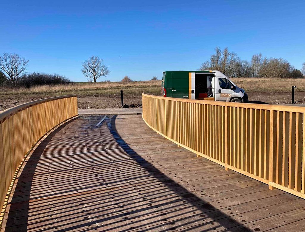 En træbro med rækværk snor sig gennem et landskab, og en hvid varevogn er parkeret i nærheden.