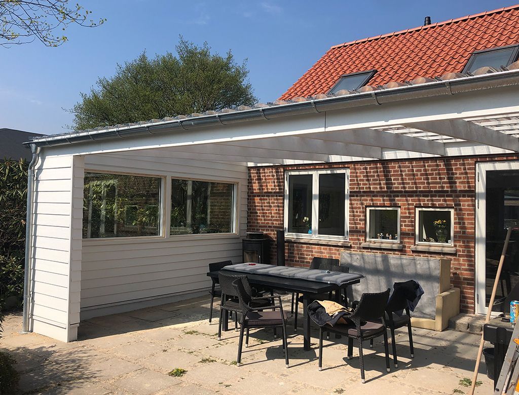 Udendørs terrasse med bord og stole under en hvidrammet pergola fastgjort til en murstensbygning.