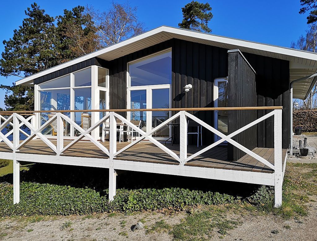 Sort hytte med hvide lister, terrasse og store vinduer, beliggende i et naturligt udendørsområde.
