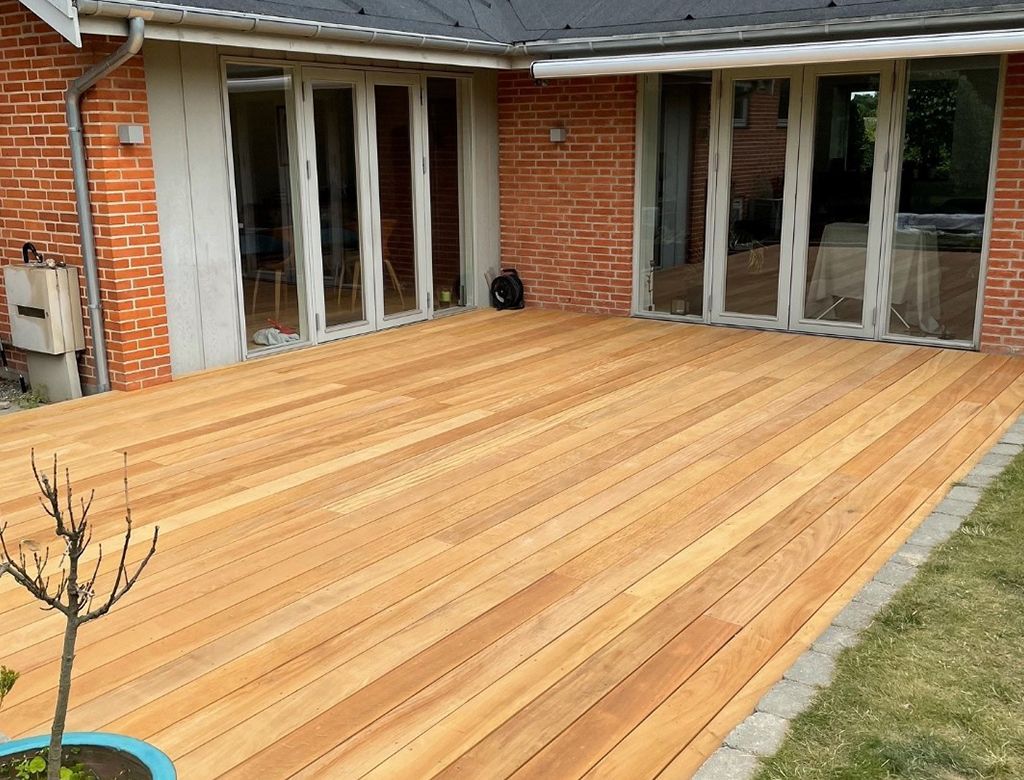 Træterrasse strækker sig fra et murstenshus med glasdøre.