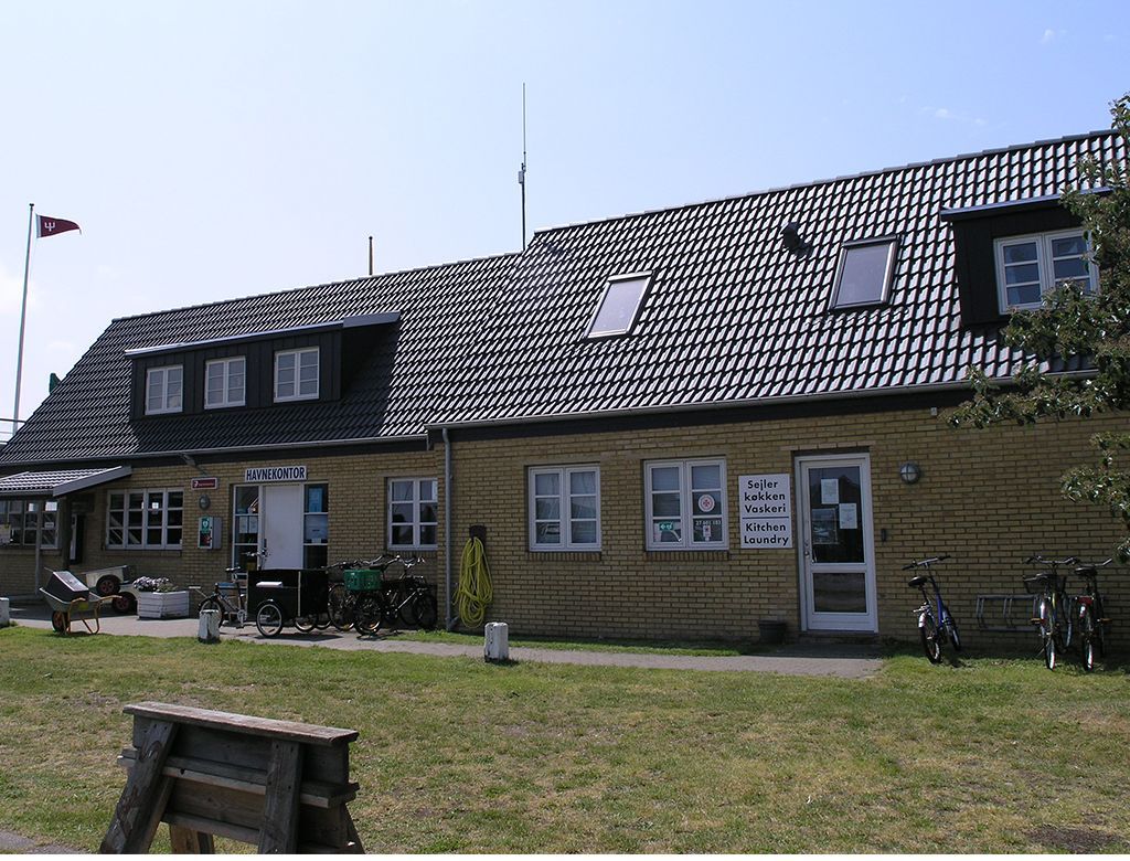 Brun murstensbygning med mørkt tegltag, kvistvinduer og et dansk flag på en stang.