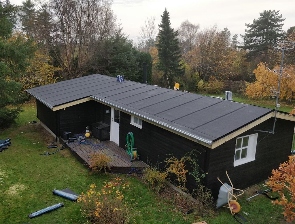 Sort hytte med nyt sort tag, terrasse og omkringliggende træer.