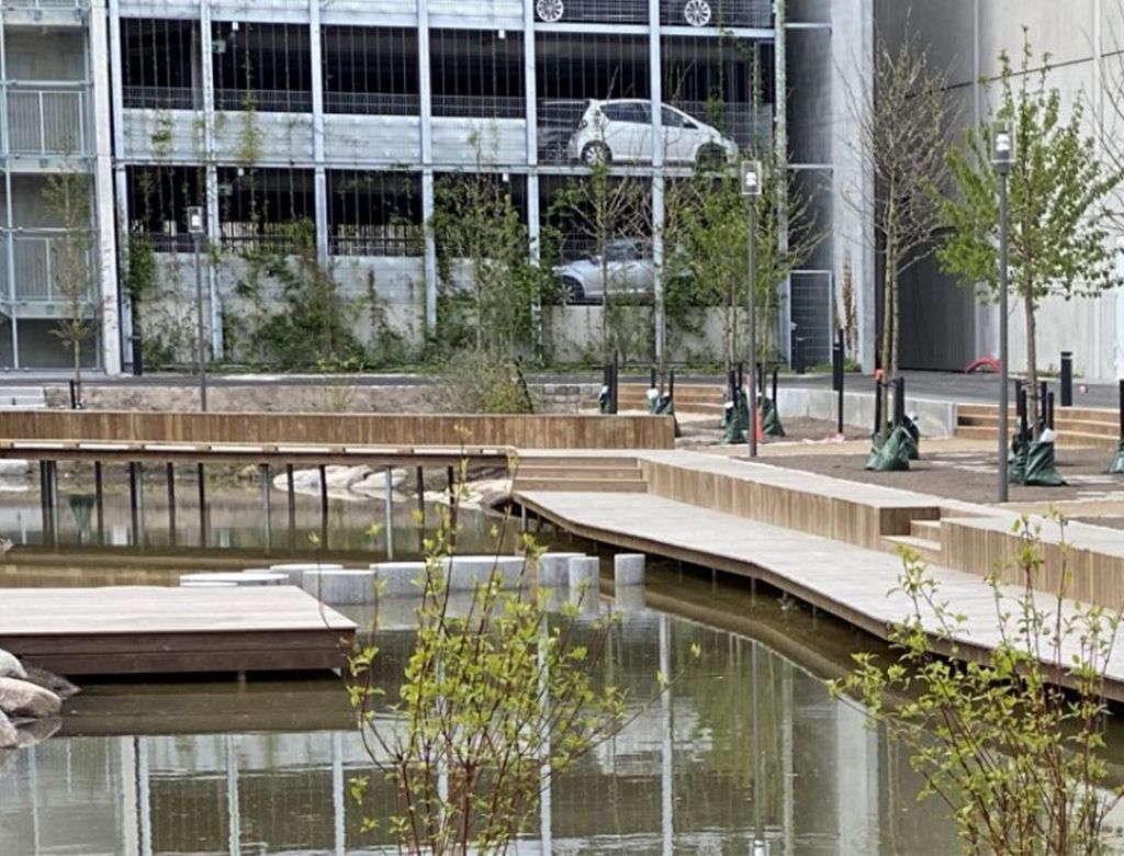 En moderne park med en dam, trægangstier og et parkeringshus i baggrunden.