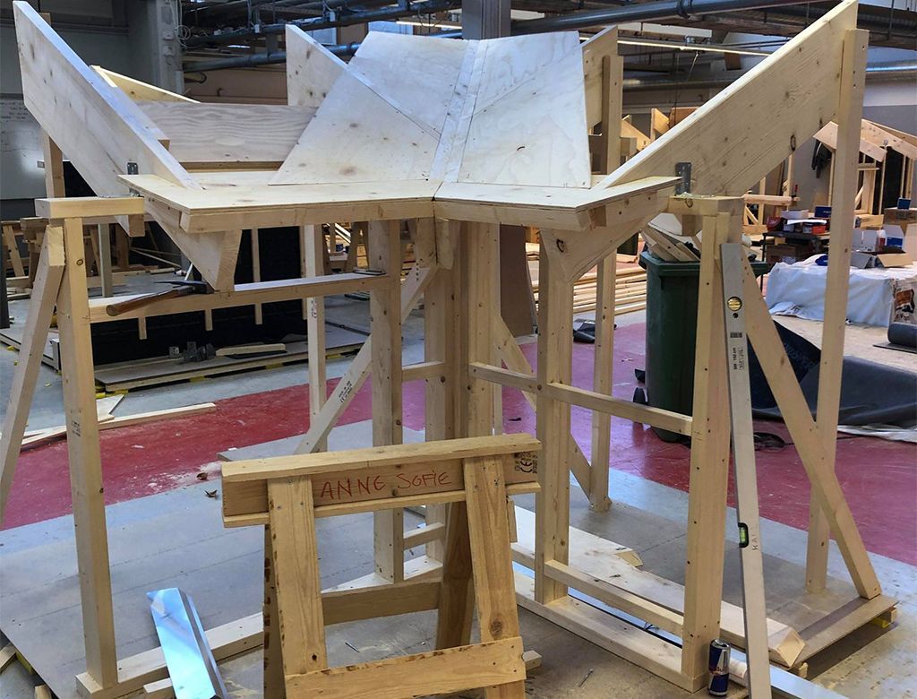 Trækonstruktion under opførelse i et værksted; lyst træ, geometriske former og støttebjælker.