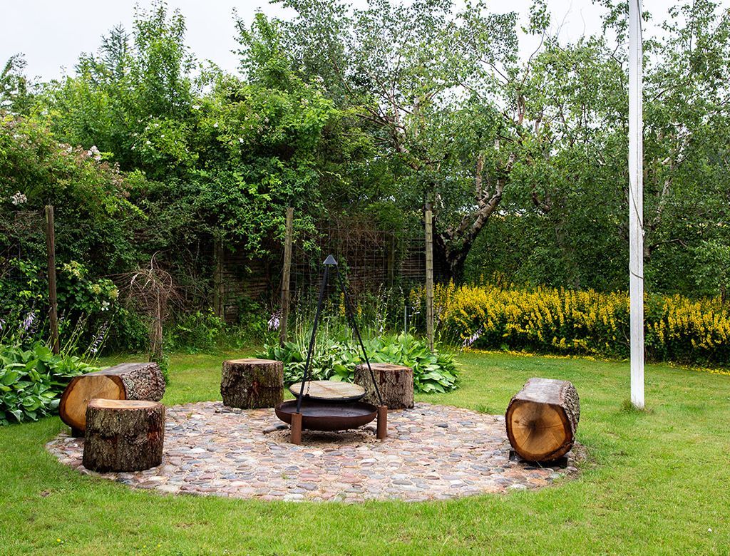 Bålsted med siddepladser i en græsklædt baghave omgivet af træer og gule blomster.