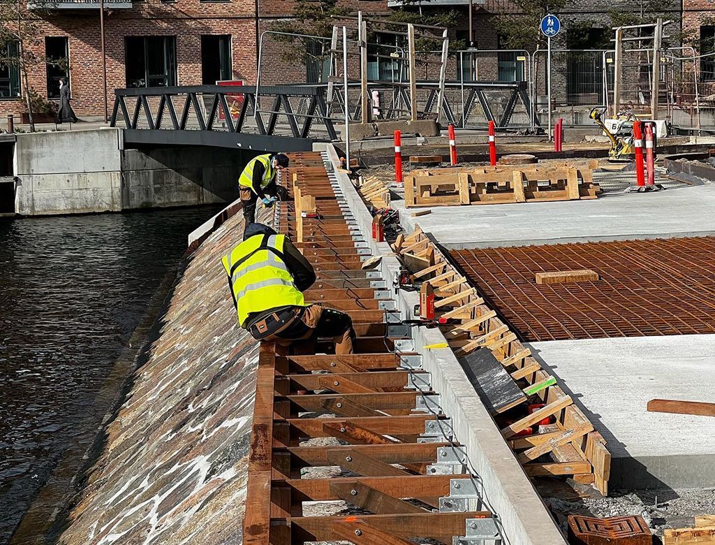 Bygningsarbejdere bygger en betonkonstruktion ved en kanal. En knæler iført sikkerhedsvest.