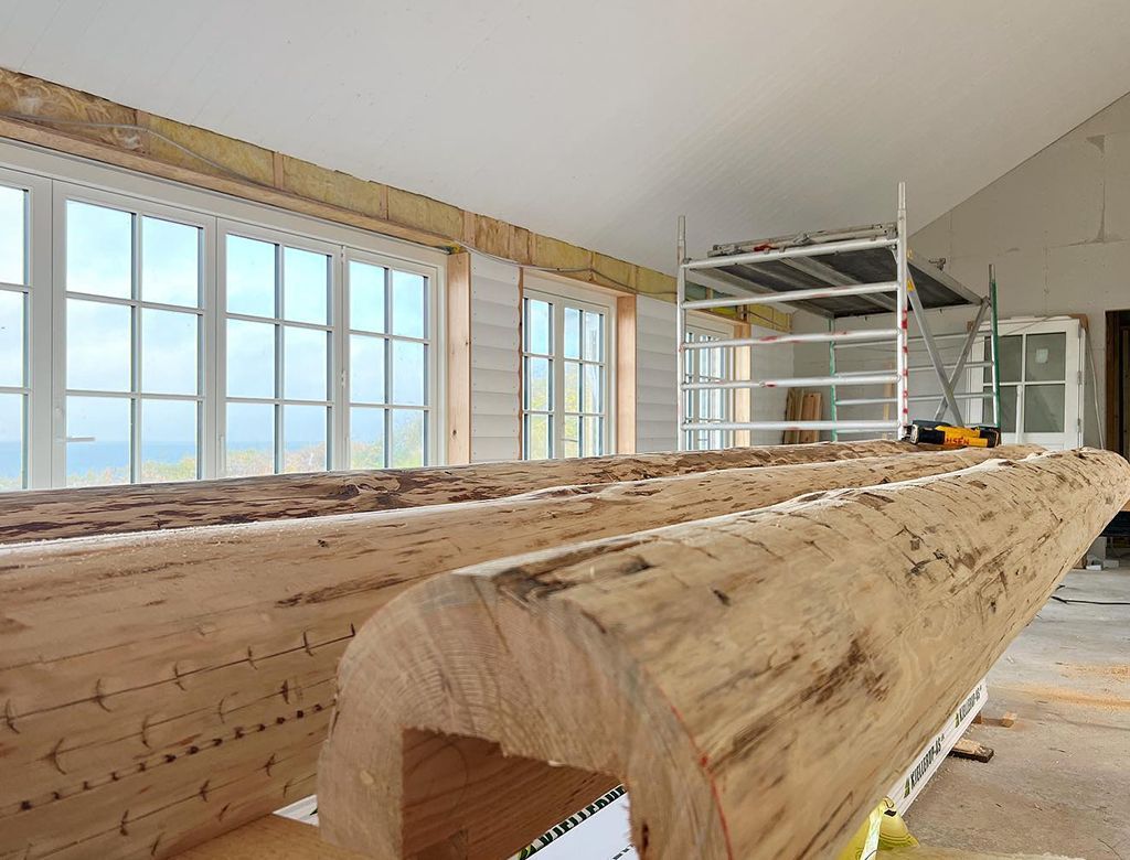 To store, ufærdige træbjælker inde i et rum med store vinduer. Byggeri i gang.
