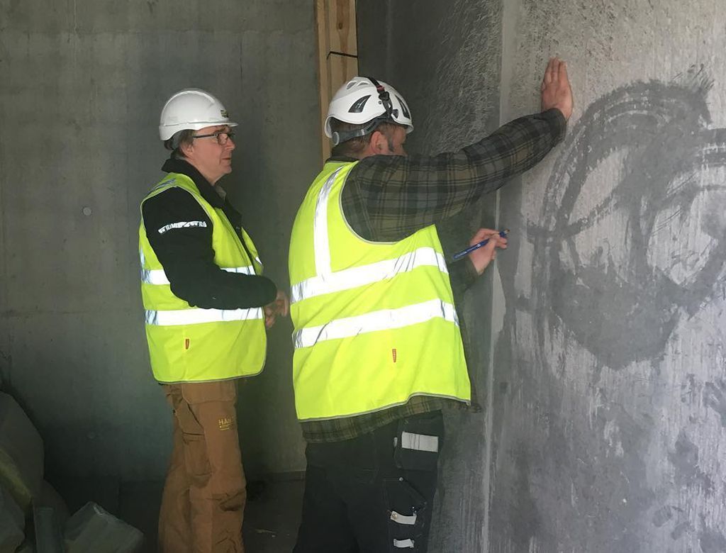 To bygningsarbejdere inspicerer en betonvæg, den ene peger, mens de indenfor bærer hjelme og sikkerhedsveste.