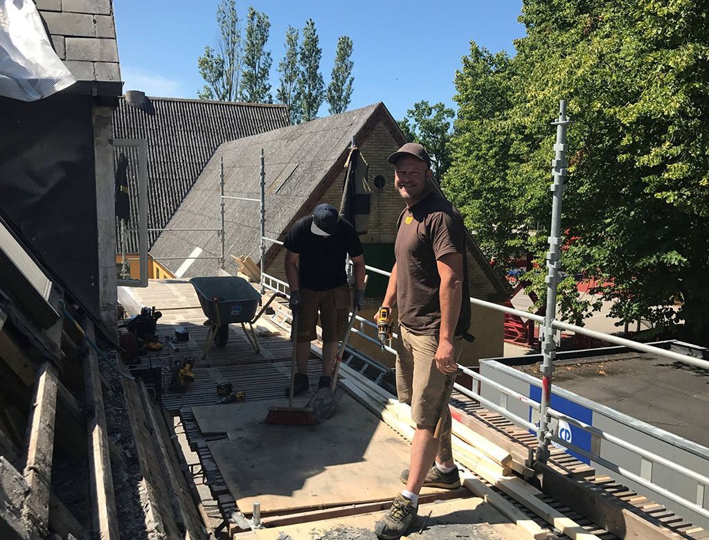 To mænd er i gang med at reparere et tag, med et delvist synligt hus i baggrunden. En mand kigger ind i kameraet.