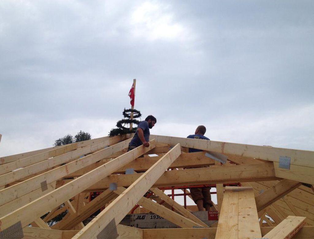 To bygningsarbejdere på en trætagramme med en dekoreret stang i baggrunden mod en overskyet himmel.