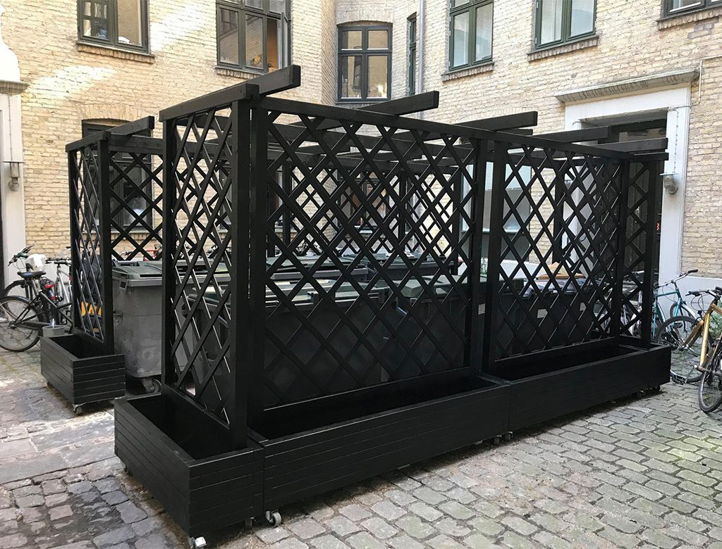 Sort udendørs siddepladsindhegning af espalier på en brostensbelagt gade; cykler parkeret i nærheden.