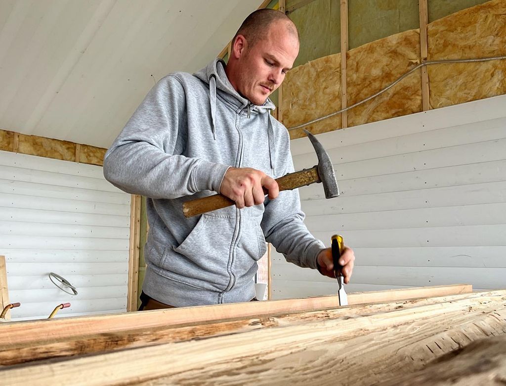 Mand skærer træ med mejsel og hammer indendørs, iført en grå hættetrøje.