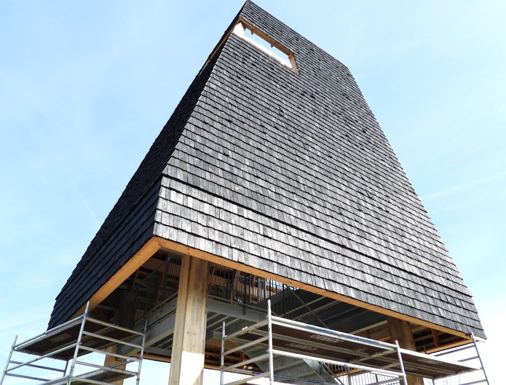 Sortbeklædt tårn med et lille vindue mod en blå himmel. Stilladser understøtter en udsigtsplatform.