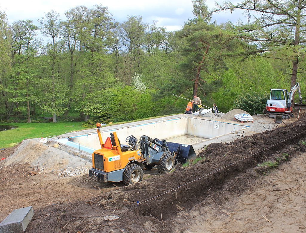 Byggeplads med tunge maskiner, pool under opførelse, træer i baggrunden.