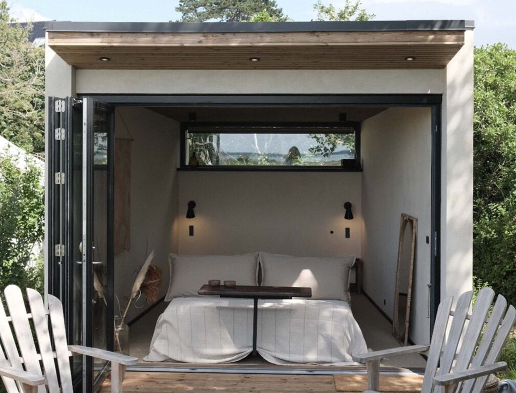 Hyggeligt udendørs soveværelse med en seng, åbne døre, to stole og udsigt til træer.