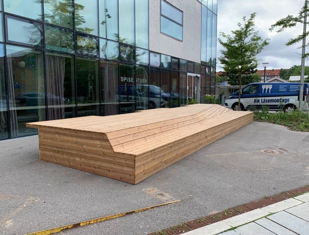 Træbænk uden for en moderne bygning, vinklet form. Brolagt område. En varevogn parkeret i baggrunden.