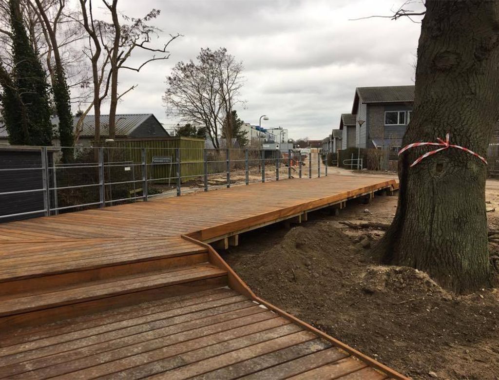En træpromenade med metalgelændere, trapper og bygninger i baggrunden. En overskyet dag.