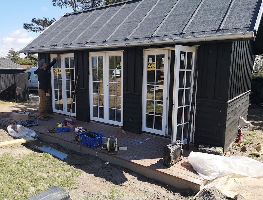 En mand står ved siden af et lille sort skur med hvidindrammede døre. Værktøj og materialer er i forgrunden.