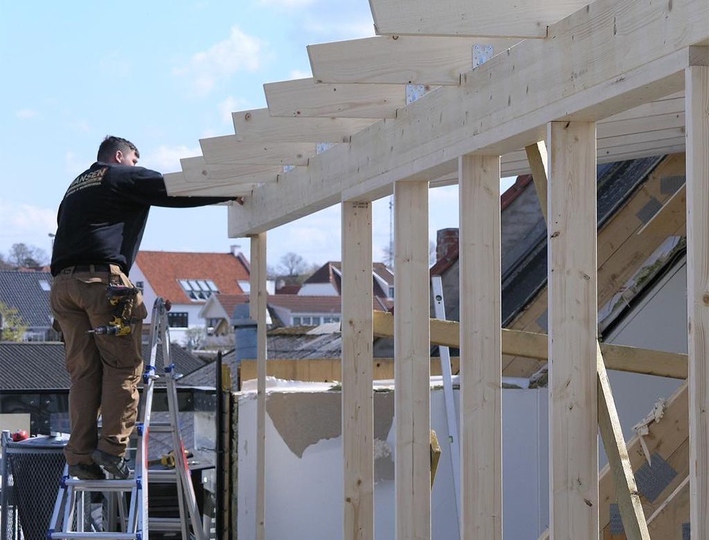 Tømrer på en stige bygger en trærammestruktur udendørs under en klar blå himmel.