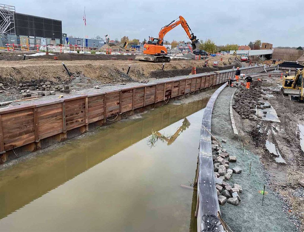 Byggeplads med en gravemaskine over en vandfyldt grøft foret med trævægge; arbejdere i nærheden.