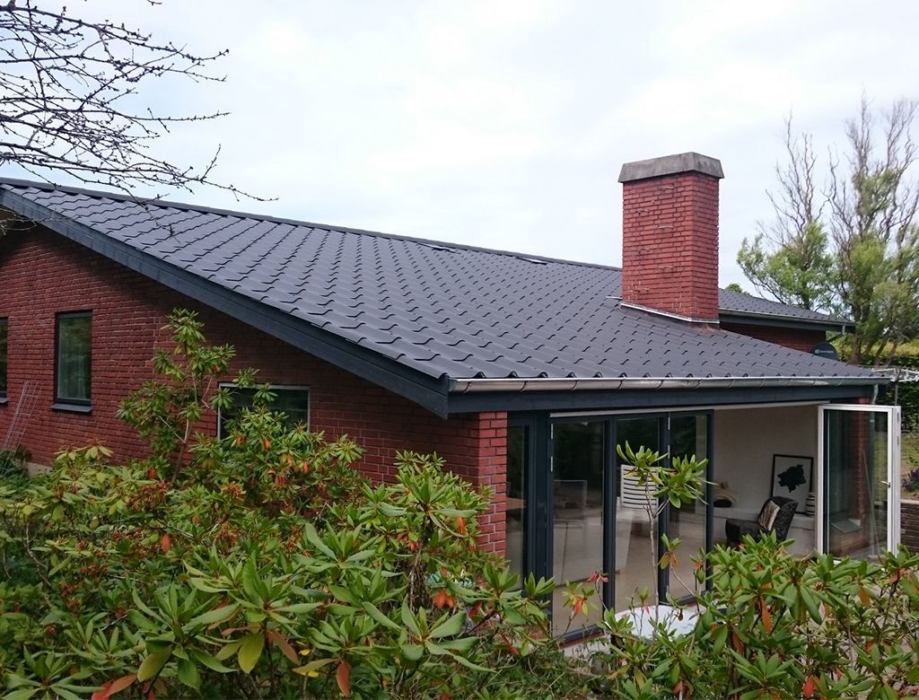 Murstenshus med mørkt tag og solrum i glas, omgivet af grønne områder og en murstensskorsten.